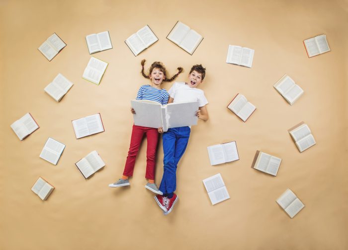 A boy and girl lying on the floor reading a large book surrounded by lots of open books