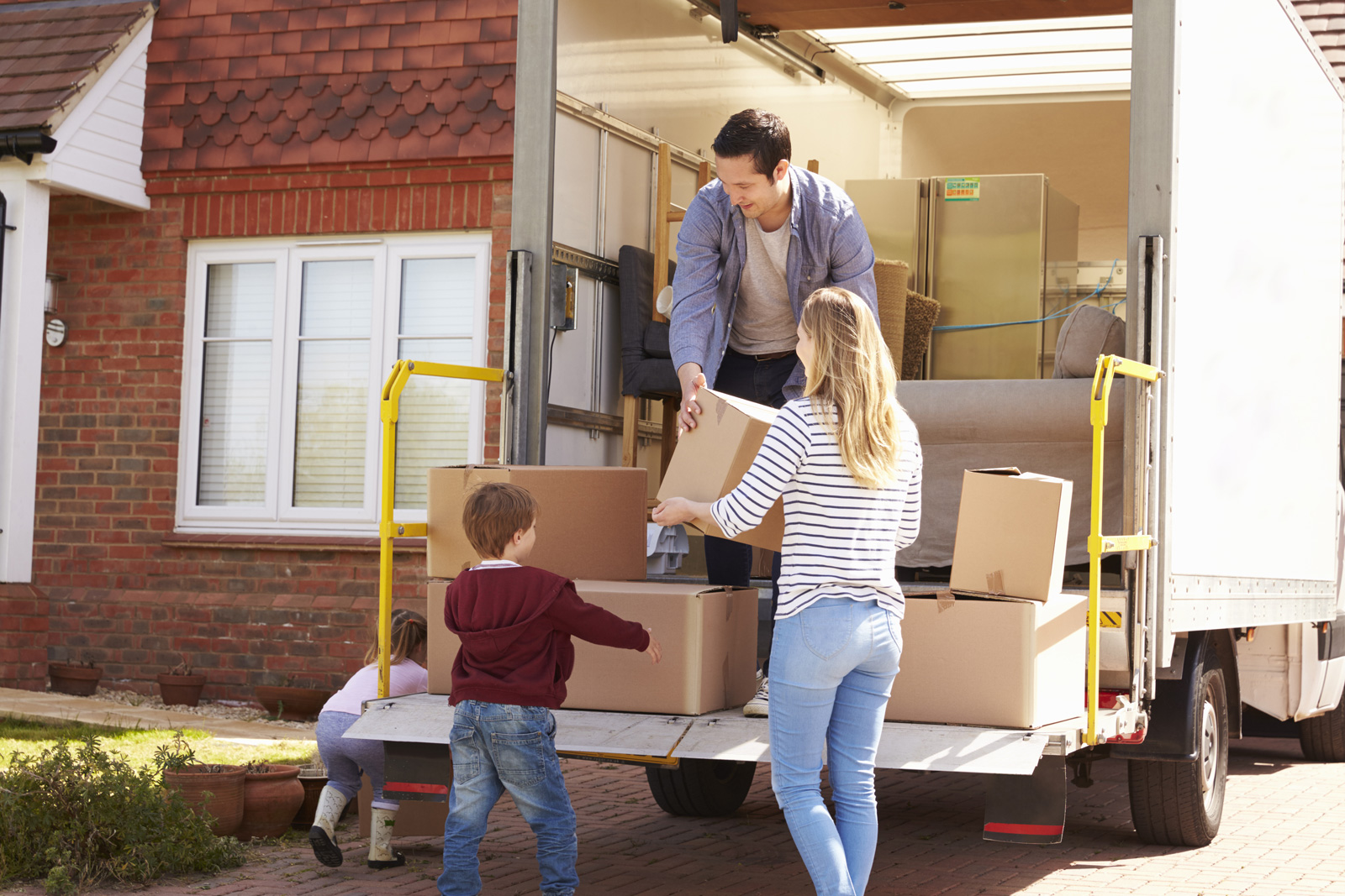 Mum and dad moving boxes out of a removals an on the driveway of their new home with their toddler son offering to help and daughter pushing a box along the drive