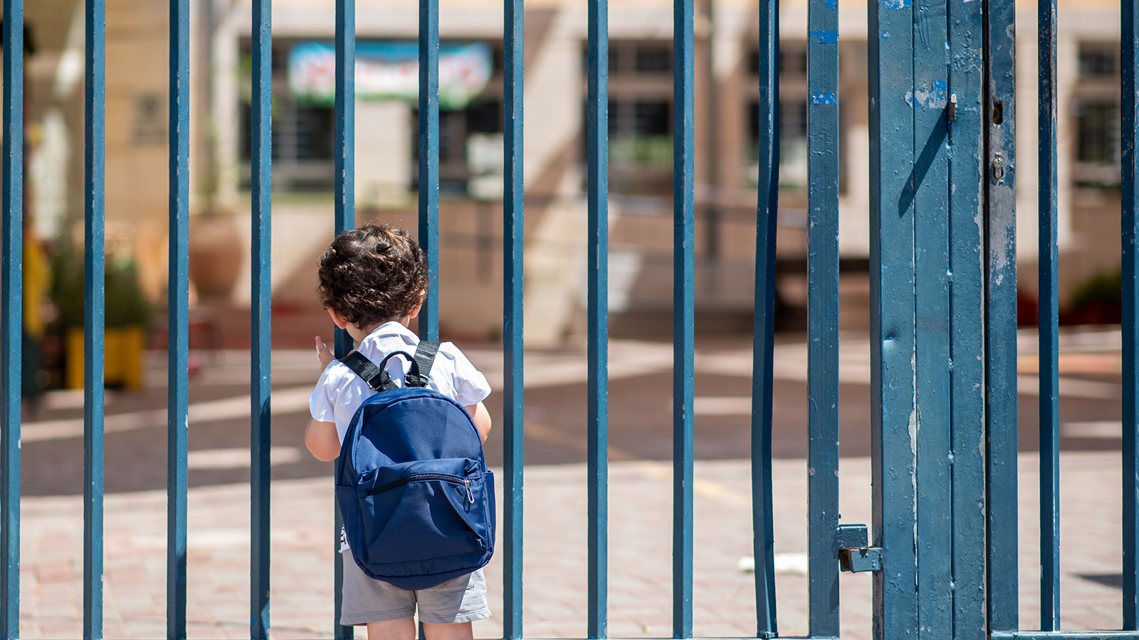 A little boy with a backpack outside some blue school gates peering inside