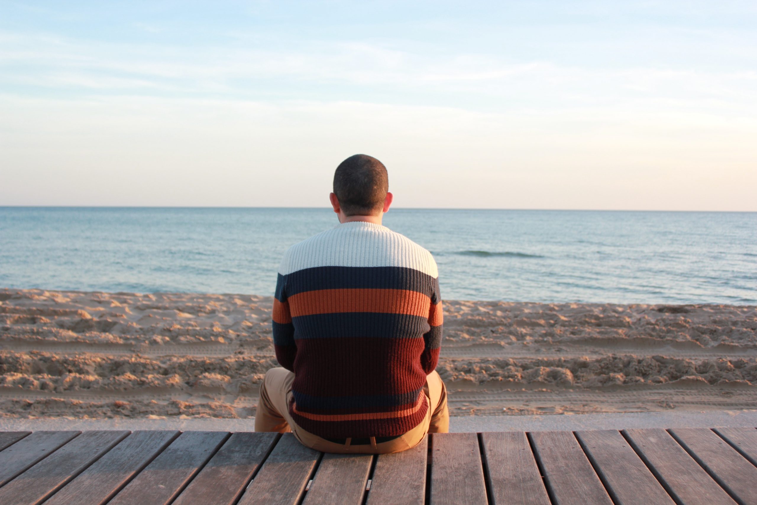A man wearing a sweater sits on a wooden dock looking out to sea