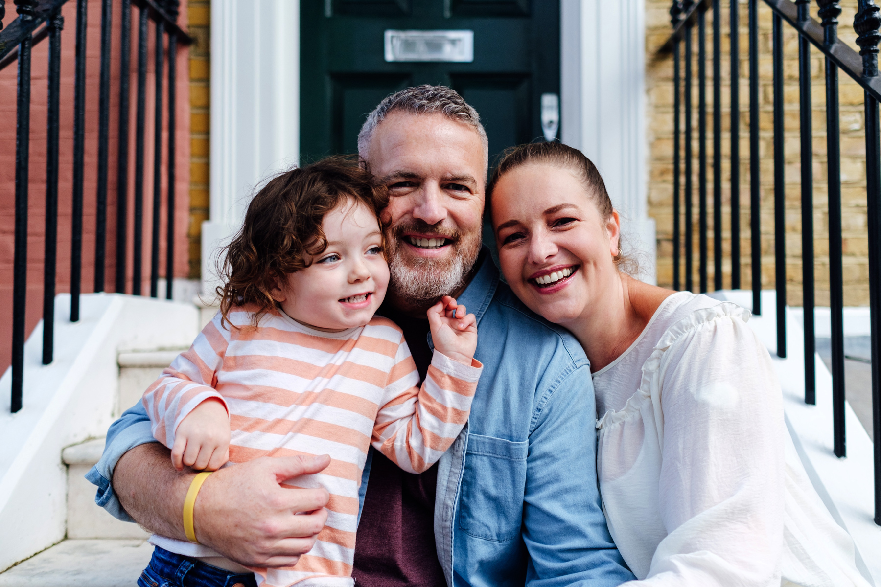 A family of three - baby girl, mum and dad - sit on the doorstep of their new home
