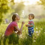 A mother blows a dandelion head with her baby son in the middle of a spring meadow.