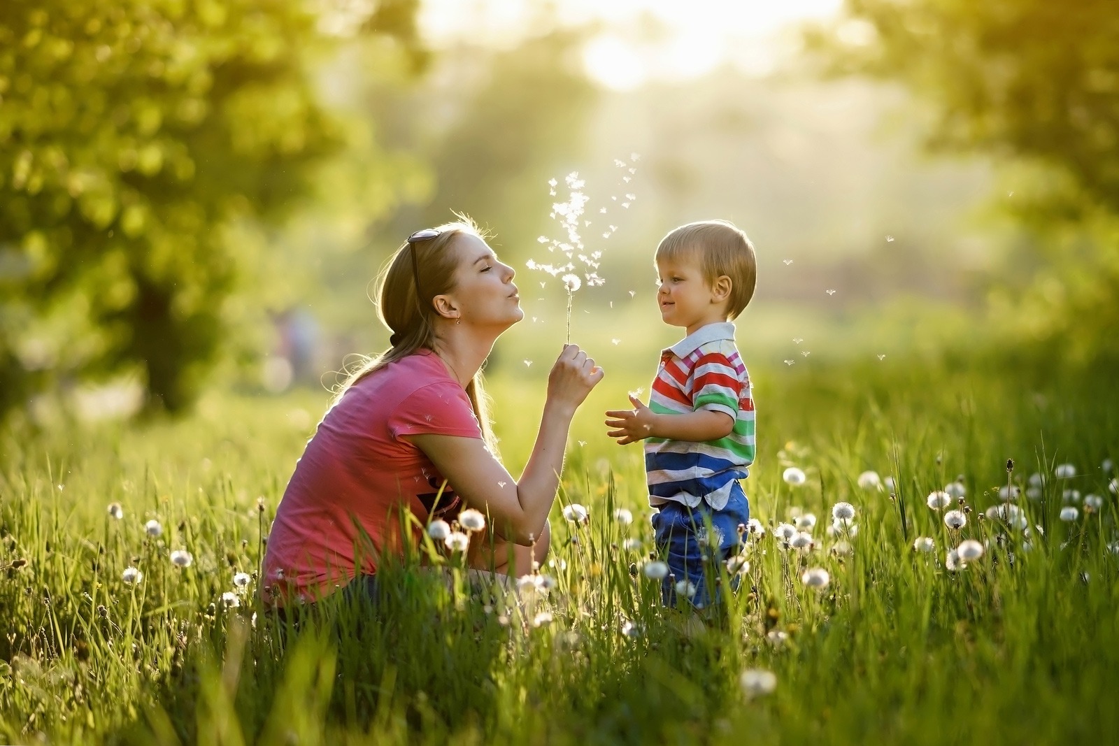 A mother blows a dandelion head with her baby son in the middle of a spring meadow.
