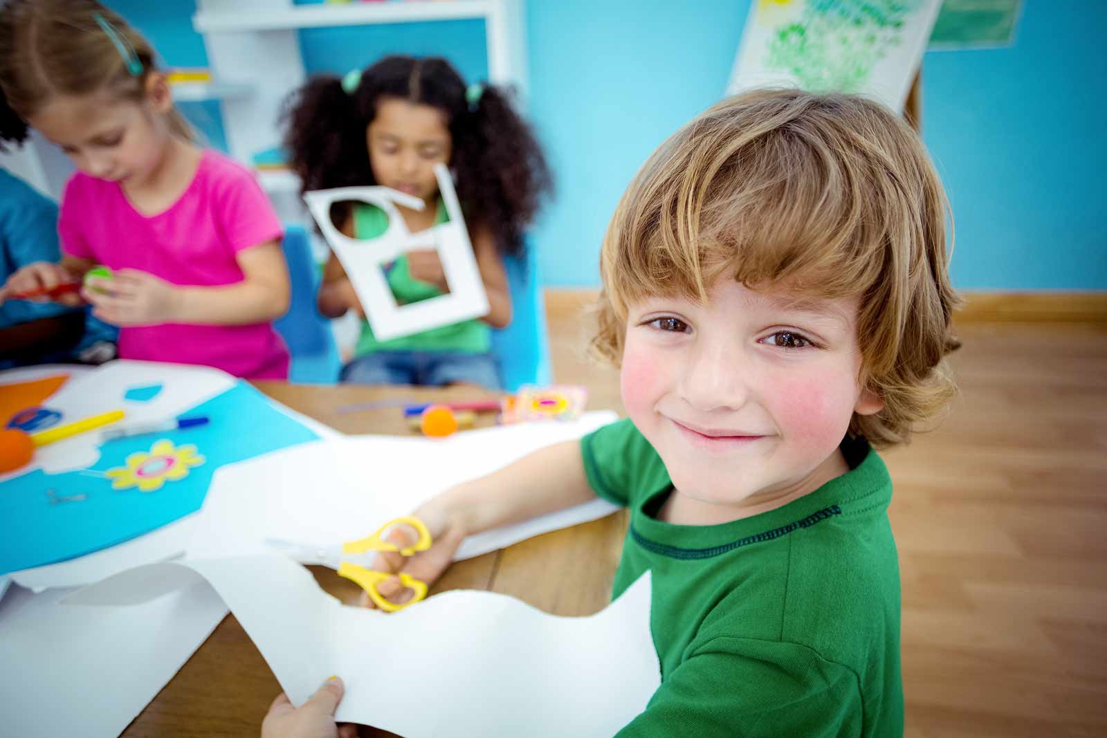 A little boy smiles at the camera while he and his two friends do arts and crafts projects.