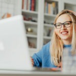 A blonde woman with funky orange over ear headphones watches a webinar on her laptop.