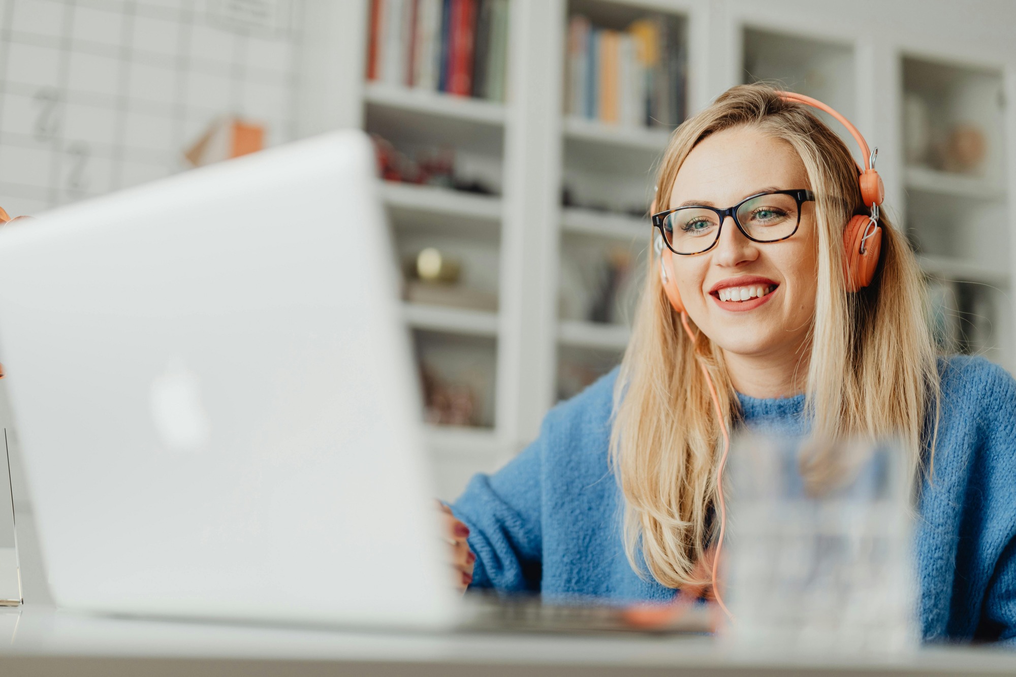 A blonde woman with funky orange over ear headphones watches a webinar on her laptop.