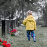 A toddler in a yellow rain jacket and wellie boots walks through a A toddler in a yellow rain jacket and wellie boots walks through a cemetary with poppies at some of the tombstones. with poppies at some of the tombstones.