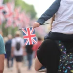 A child sits on someone's shoulder holding a Union Jack flag, as they walk down a flag lined street.