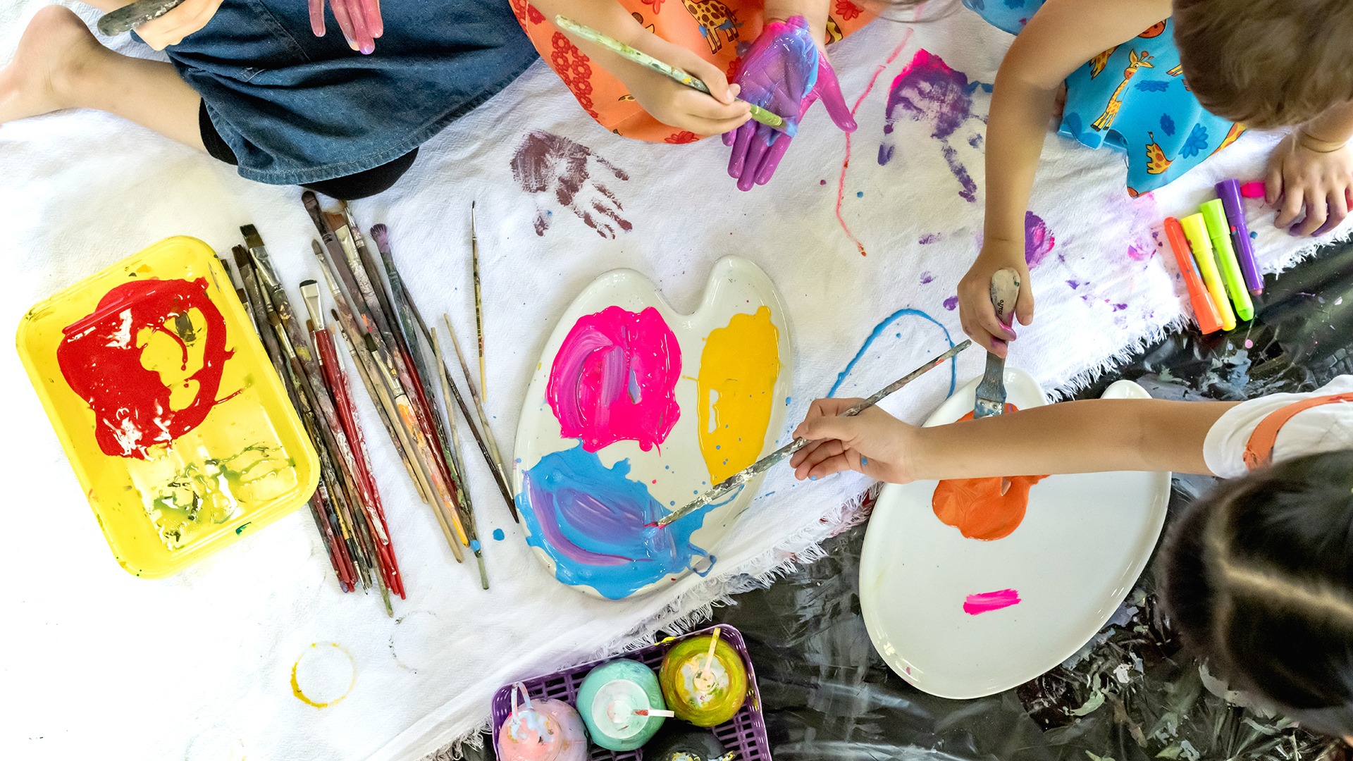 A close-up, overhead shot of several children's hands painting with bright colors on a white dropcloth. There are paintbrushes, two palettes with pink, yellow, blue, and orange paint, and various paint smears and handprints visible.